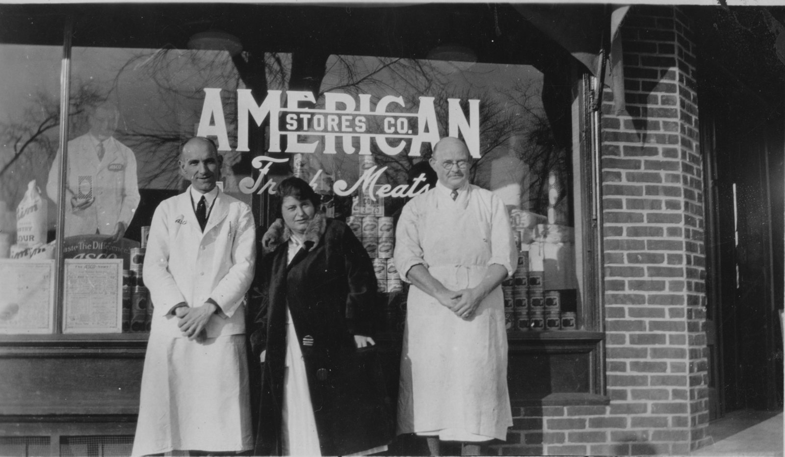 Three staff in white aprons and a woman in a dark coat stand at the store window, tall stacks of canned goods behind the glass and American Stores Co. lettered above.