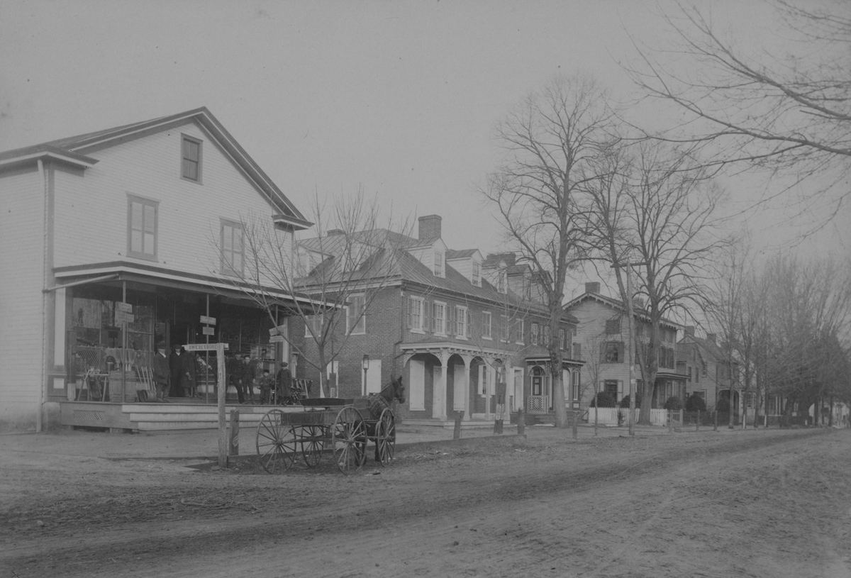 An unpaved Main Street lined with brick and frame buildings, a horse-drawn cart waiting at the corner store's porch.