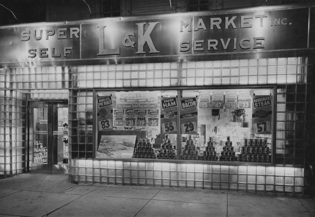 The L&K Market storefront in glass block and steel, Super Self Service lettered across the front, the window stacked with can displays.