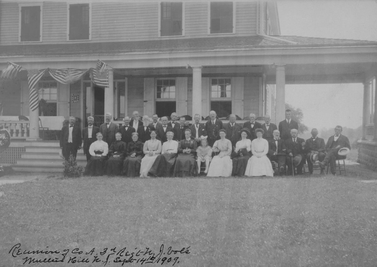 About forty veterans and their wives posed on a lawn before a porch, the women seated in long white dresses.