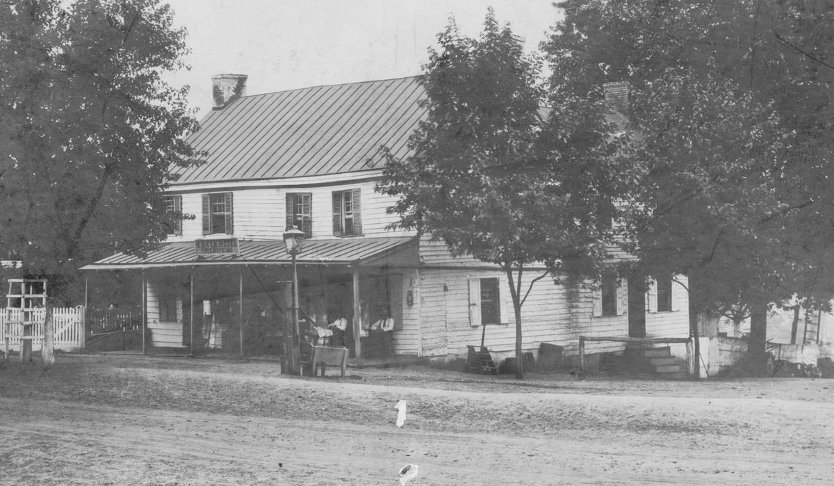 The long two-story hotel behind bare trees, figures on the porch, the street unpaved in front.
