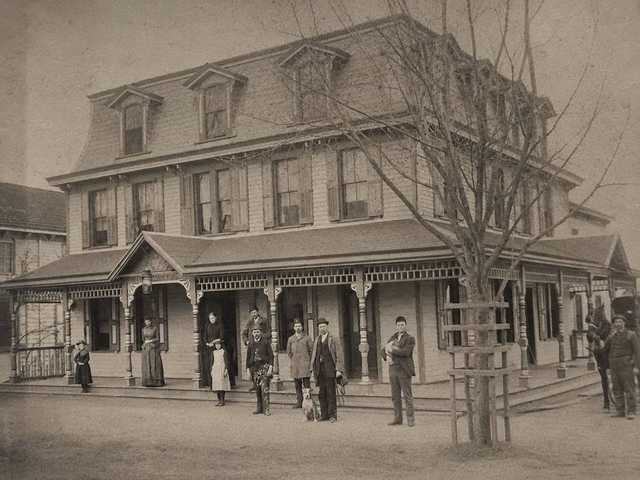 Family and staff lined up along the hotel porch rail, a young staked tree at the curb.