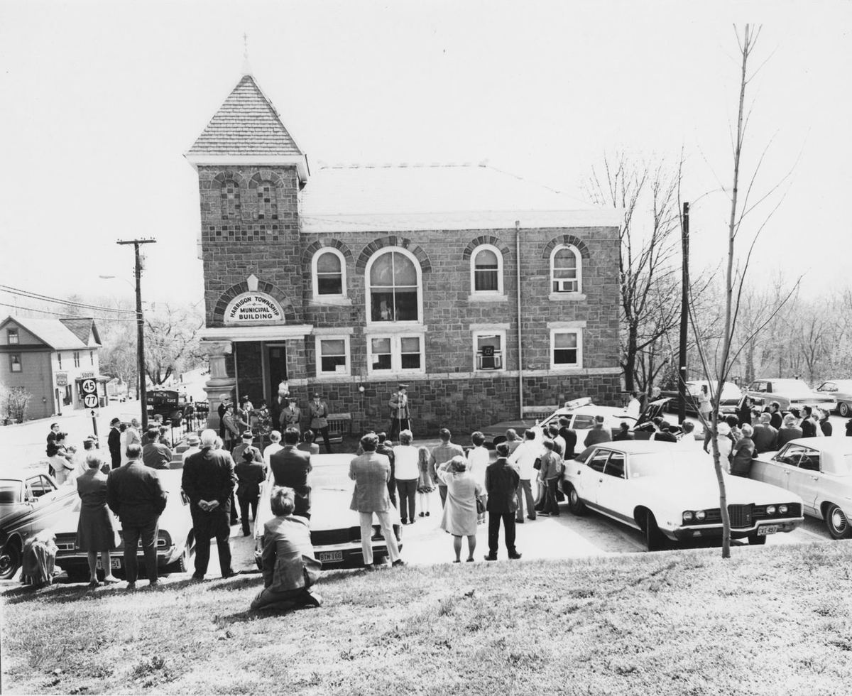 A crowd on the old bank's lawn around parked cars, children seated on the grass in front.