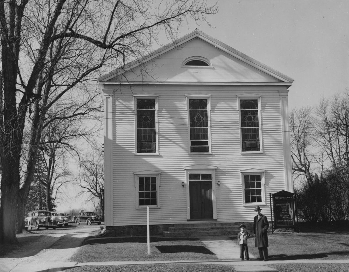 The white clapboard church face-on with tall arched windows, a man and child by the sign at the sidewalk.