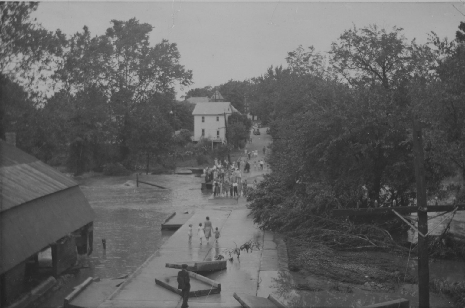 Floodwater covering the road by the mill, people wading and a rowboat in mid-street.