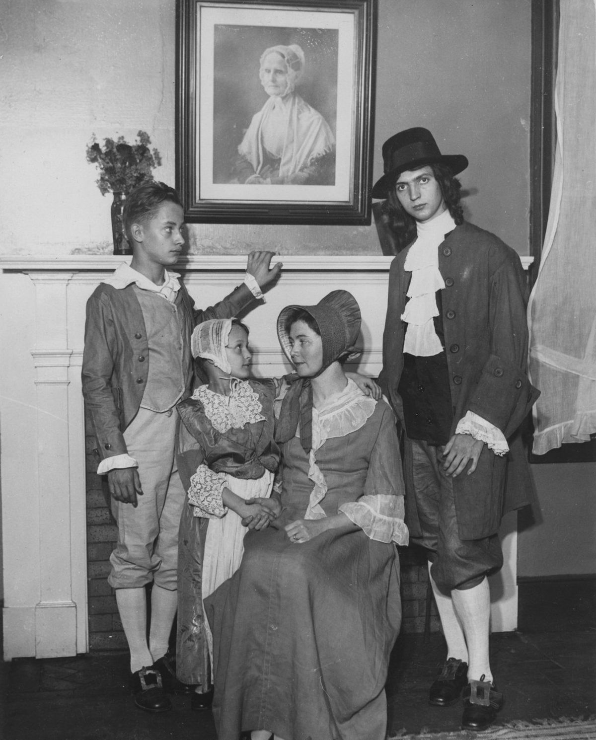 Four children in Quaker costume, broad hats and bonnets, posed beneath a framed portrait.