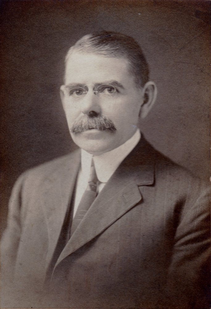 Studio portrait of a mustached man in a dark suit and glasses.