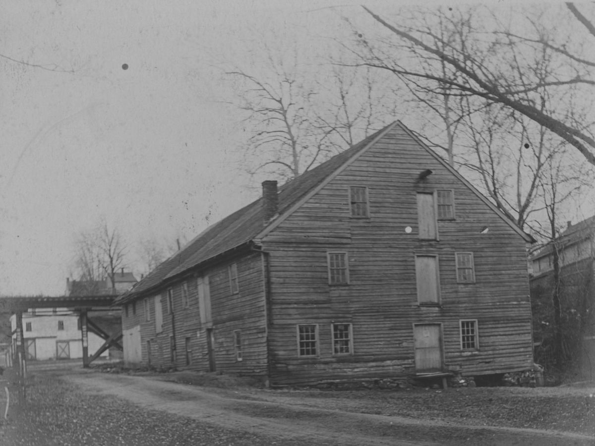 The tall three-story frame gristmill beside the road, its siding weathered, loading doors up the gable.