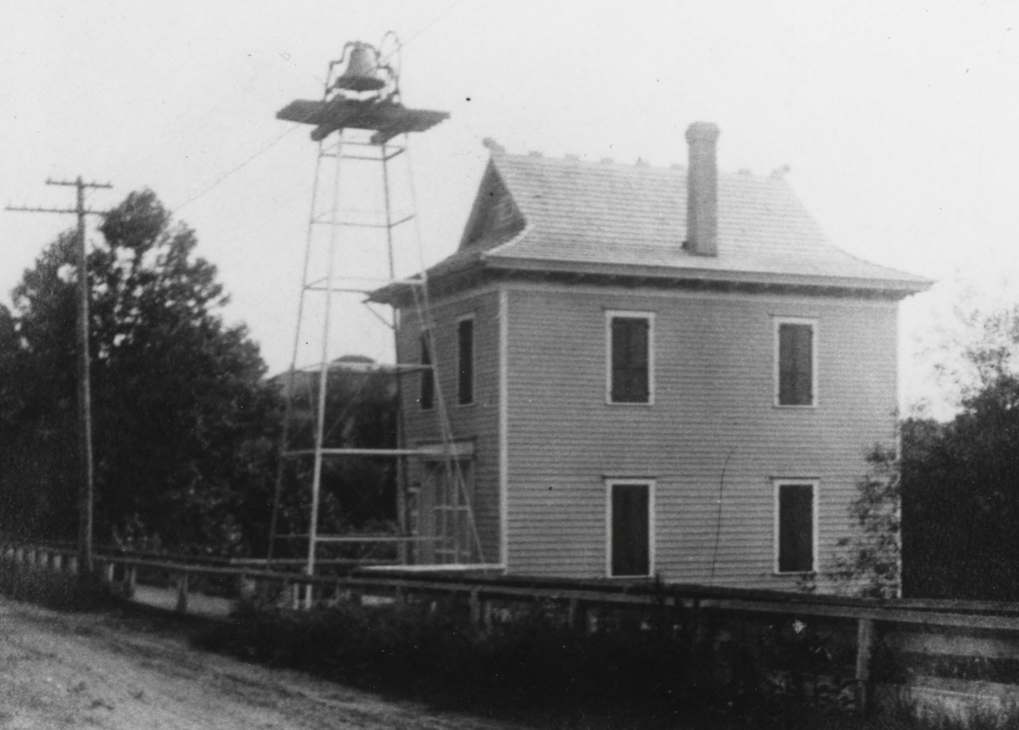 The two-story frame firehouse with its fire bell raised on a steel tower beside it.
