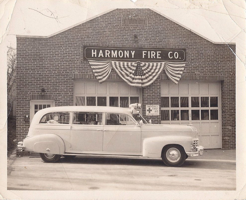 The brick fire station hung with flag bunting, Harmony Fire Co. lettered above the bays, an ambulance backed into an open bay, and a Red Cross Emergency First Aid sign beside the door.