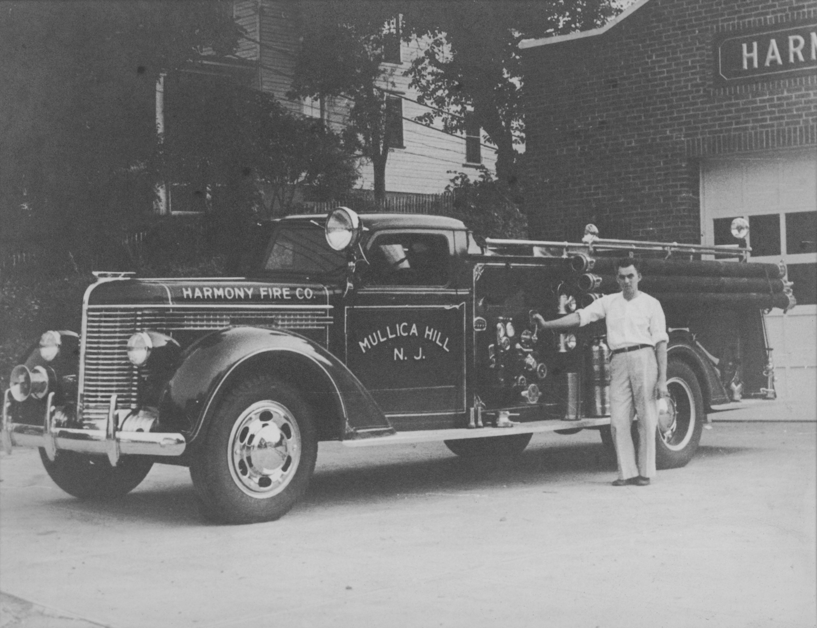 A Harmony Fire Company pumper parked outside the station, Mullica Hill N.J. lettered on its side, hose and hand rails along its body.