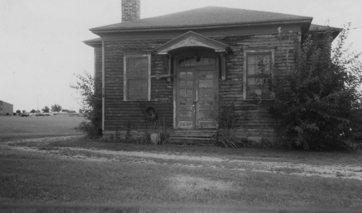 The one-room schoolhouse with a small entry hood, its doors boarded, open fields behind.