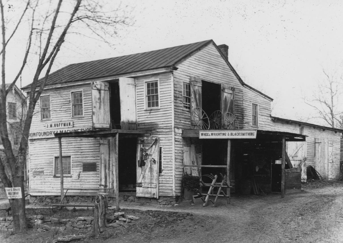 The two-story shop building with signboards on its face for wheelwrighting and blacksmithing, a wheel leaning at the open bay.