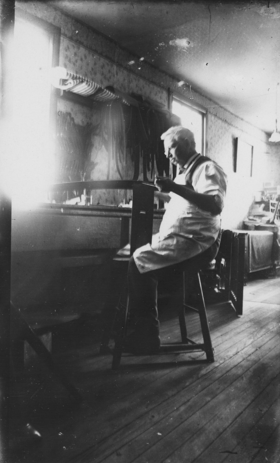 A man at work on a stool by the shop window, straps and tools on the dark walls around him.