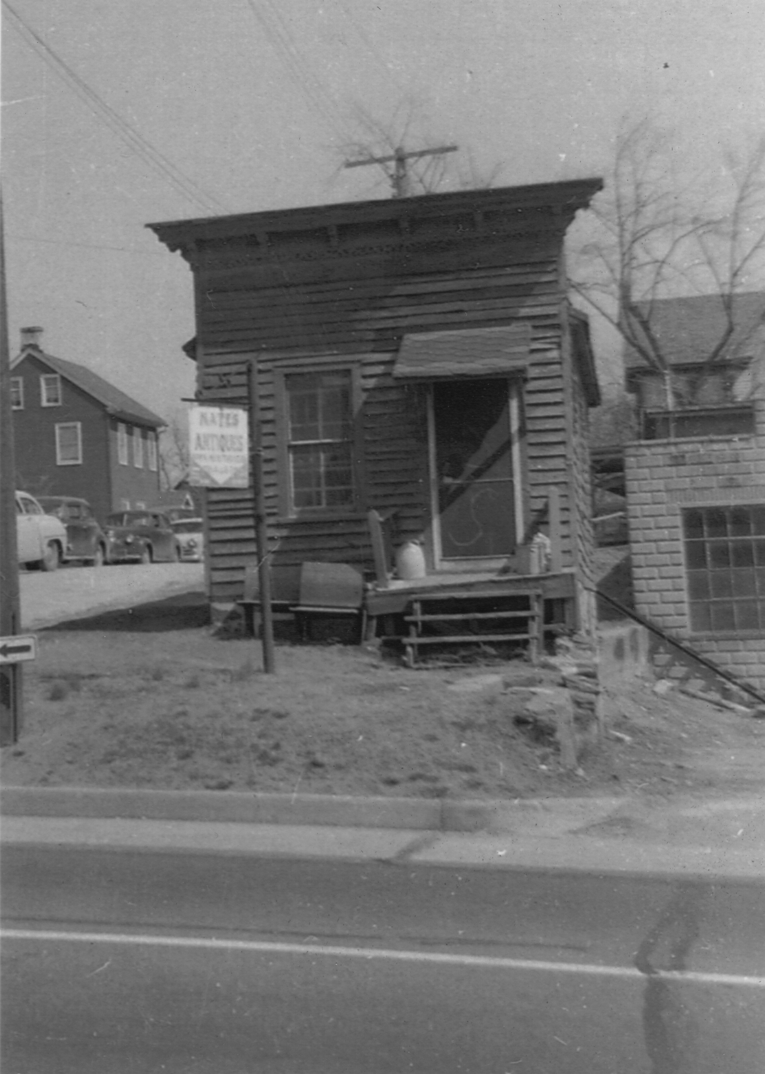 The small storefront with a hanging Antiques sign beside the window, older cars parked across the street and a masonry garage beside it.