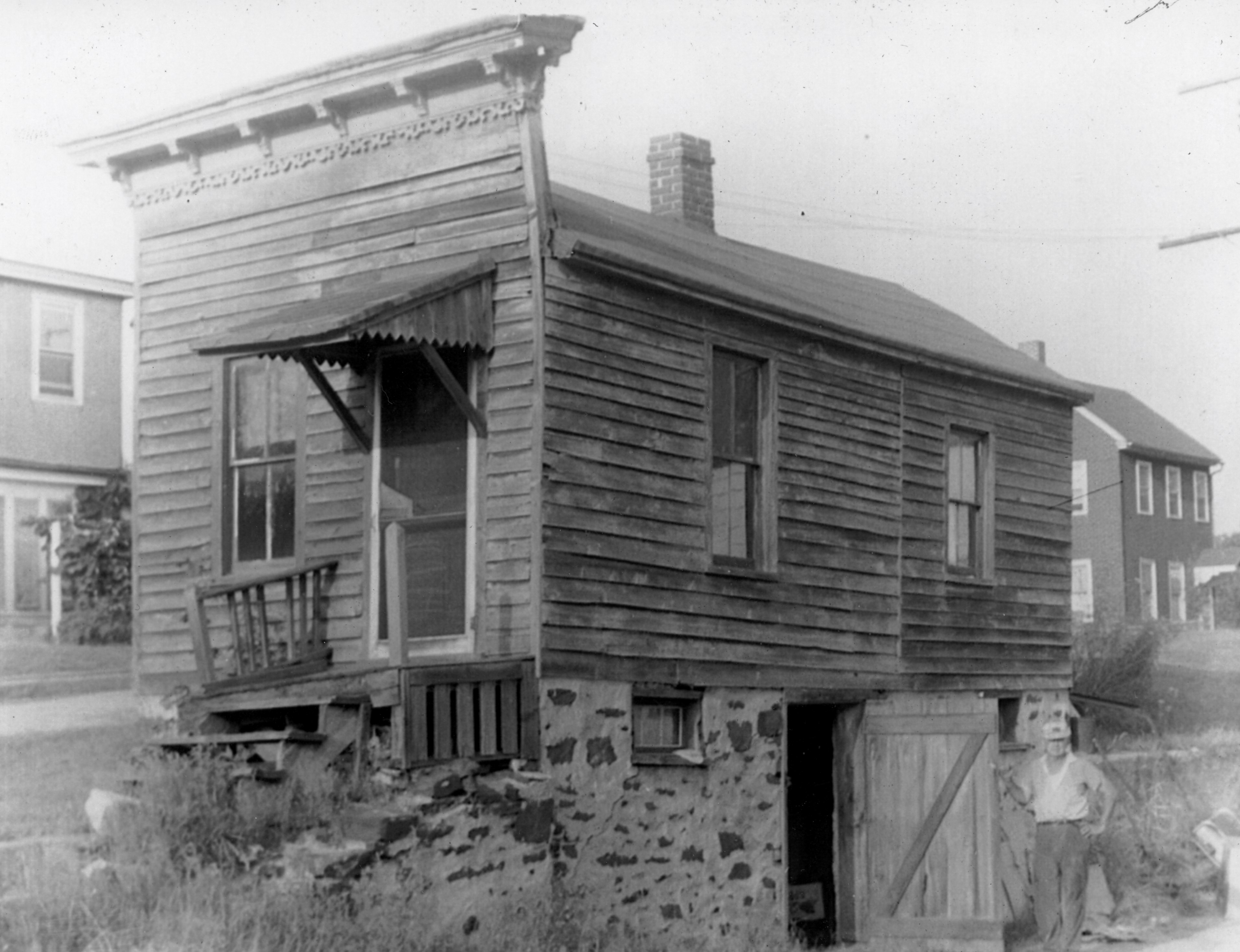 The derelict narrow frame building with a bracketed storefront cornice, standing over a raised stone basement; a man stands at the open basement door on the side street.