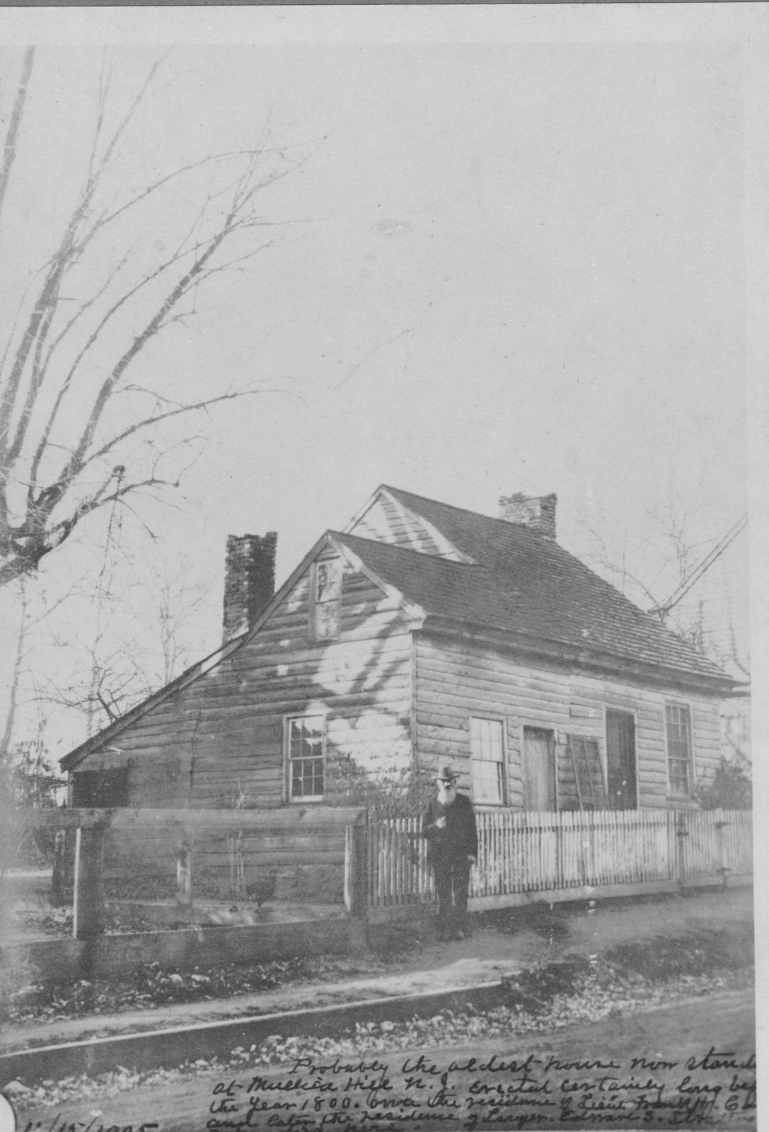 An old print of the low one-and-a-half-story house with a brick chimney at its end, a figure standing at the fence.