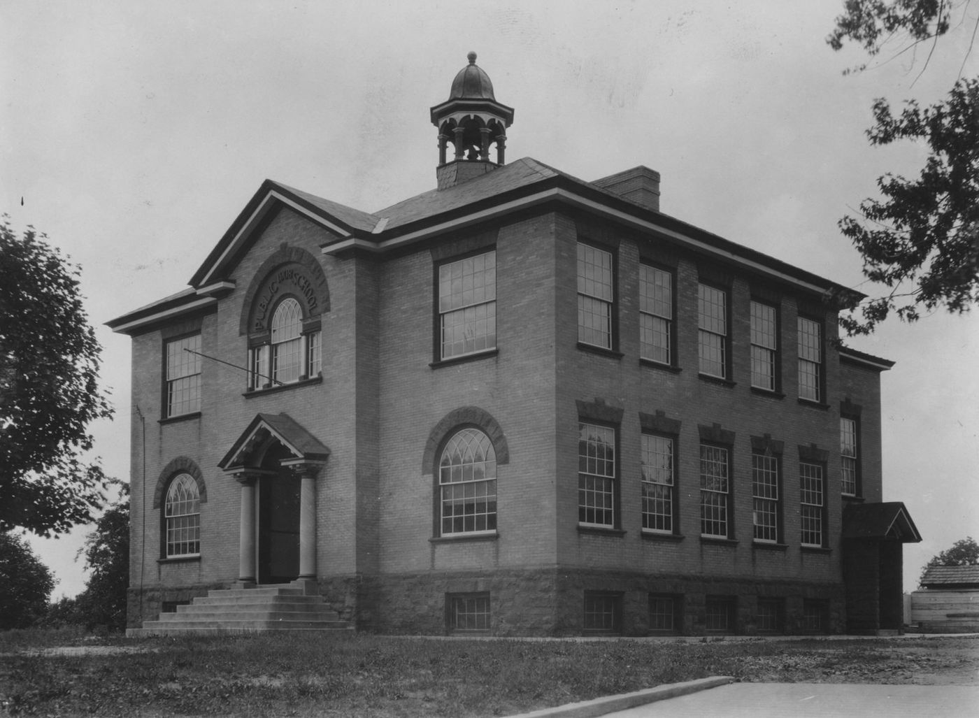 The square two-story stuccoed school under a hipped roof, an arched window in its front gable and a cupola on top.