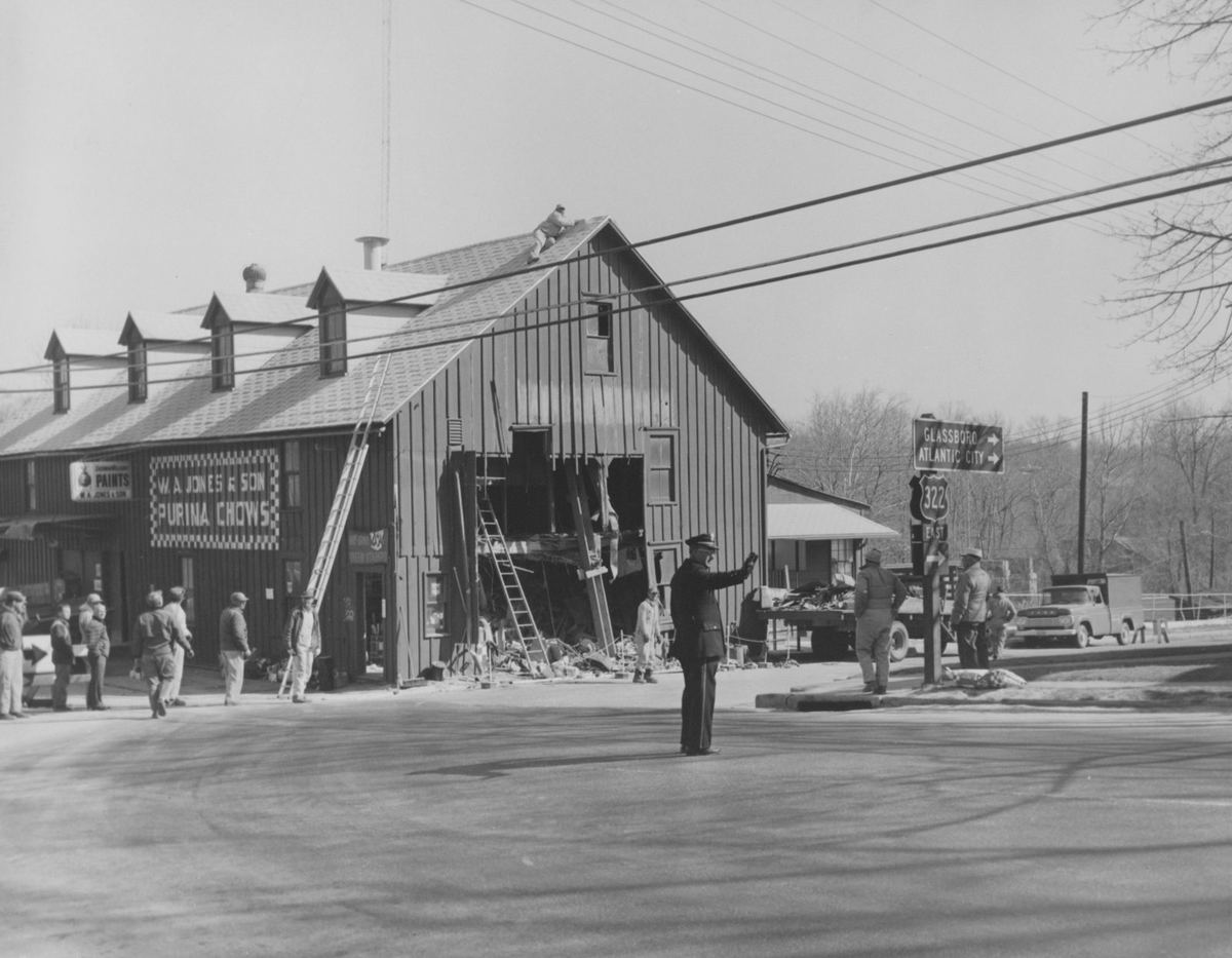 Workmen and a boom truck at the tall mill building, the street closed around the damaged corner.