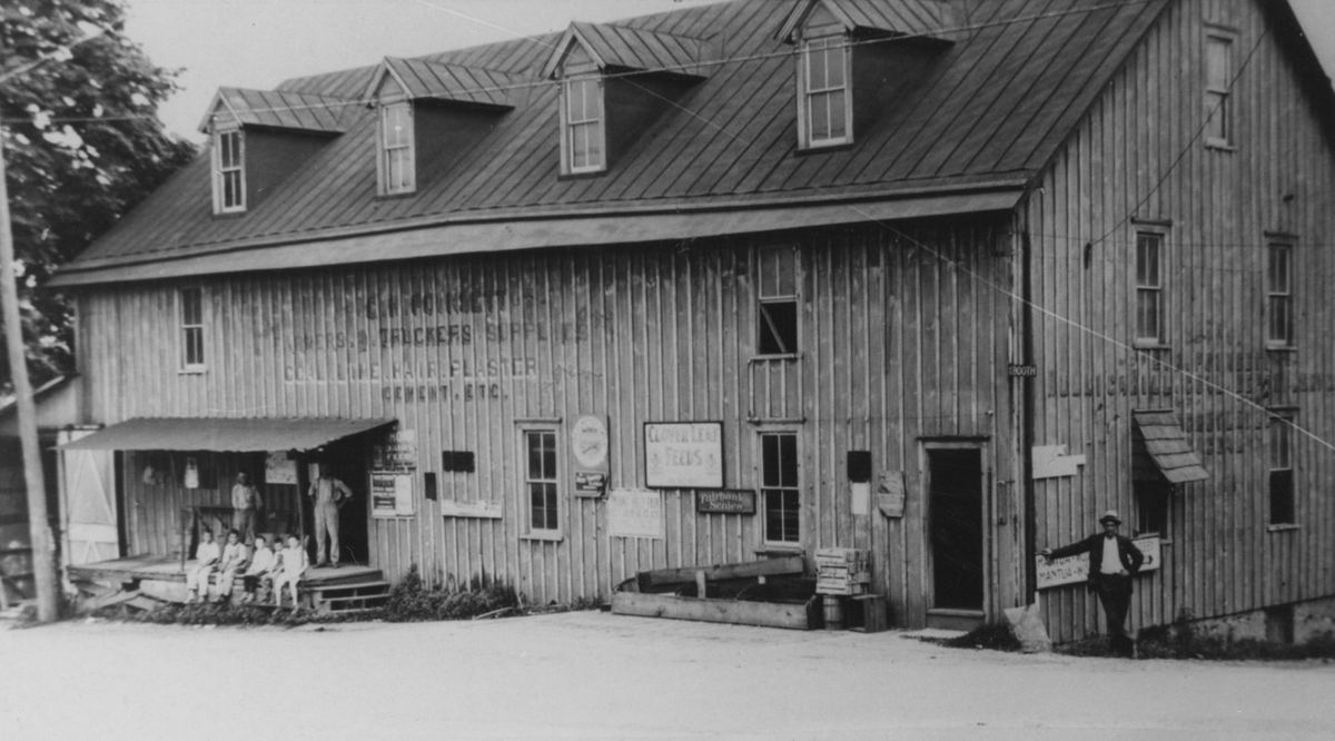 The broad three-story feed store with painted signs across its boards, a group seated on the loading porch and a man standing at right.