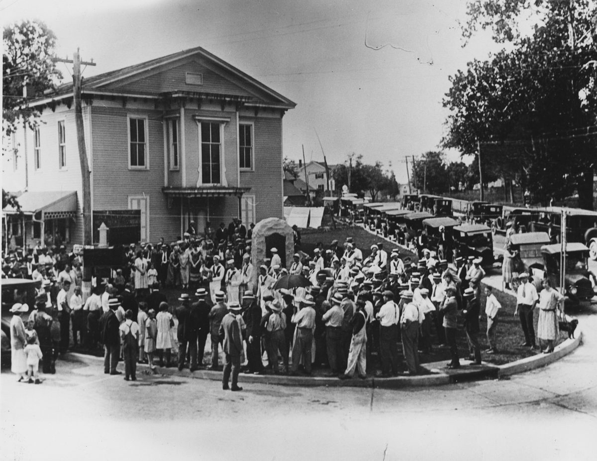 A dense crowd filling the street corner before the hall, cars parked along the curb.