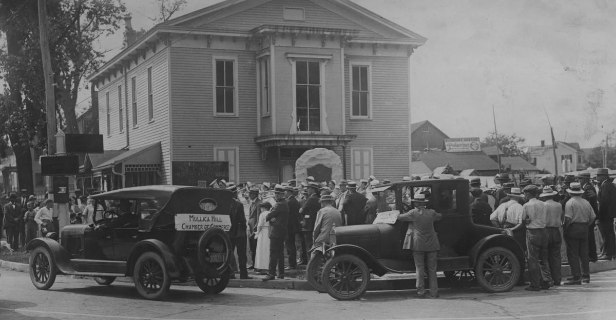 Men in hats crowded around two open touring cars before the hall's porch.