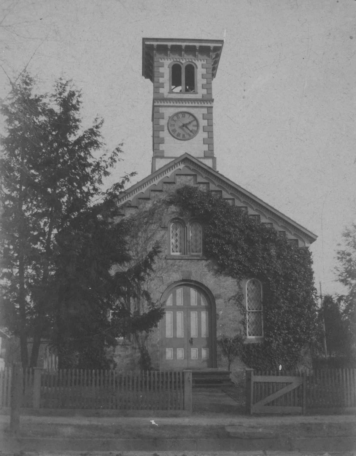 The stone church behind a picket fence, ivy grown up its square clock tower.