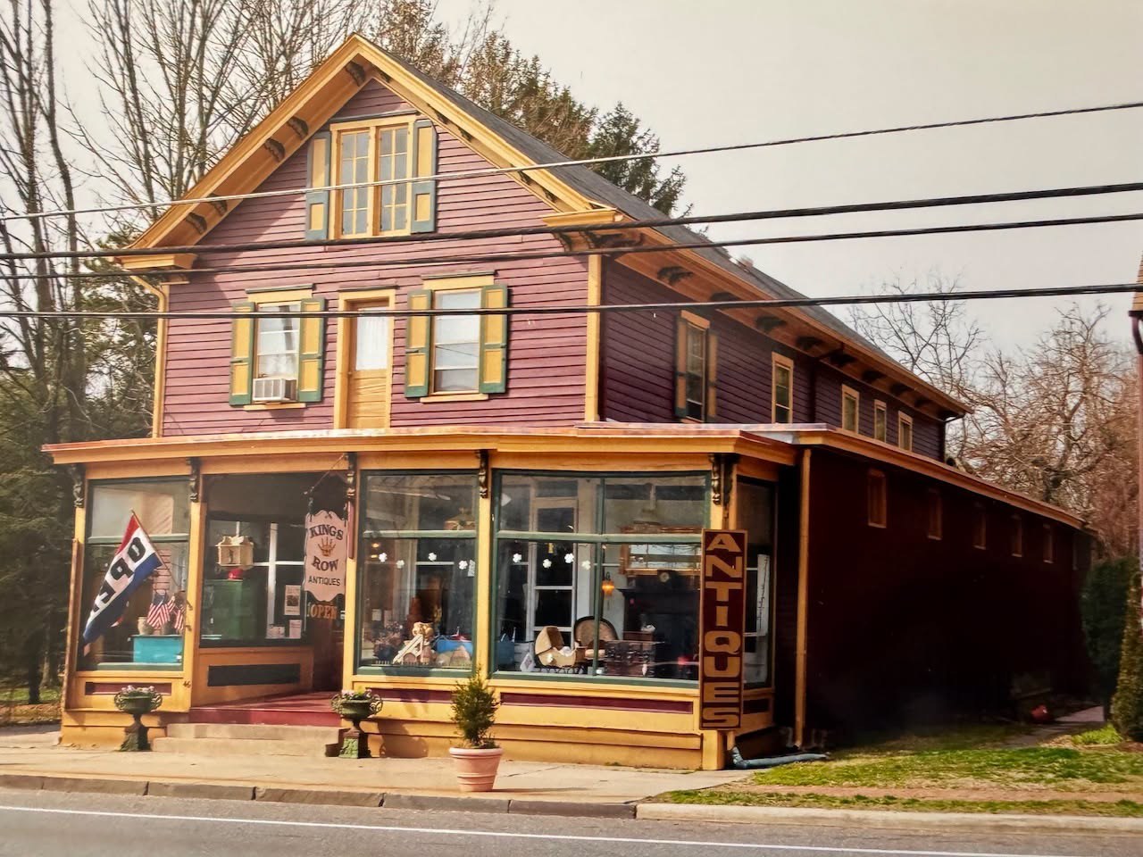 The tall gabled store painted in two colors, Kings Row Antiques signed over the porch and a vertical Antiques sign beside its display windows.