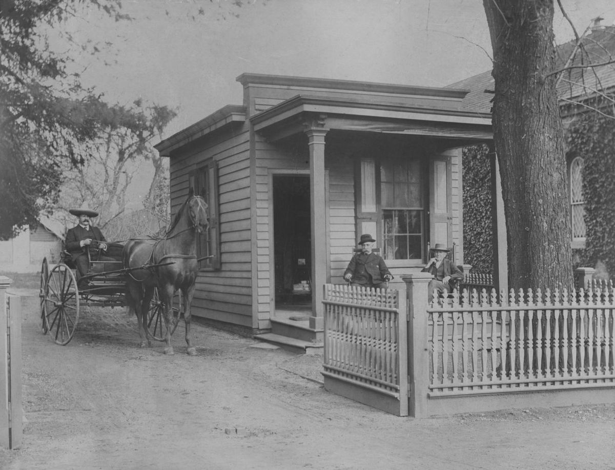 A horse and buggy at the picket fence of the little office wing, two figures by its porch beside the main house.