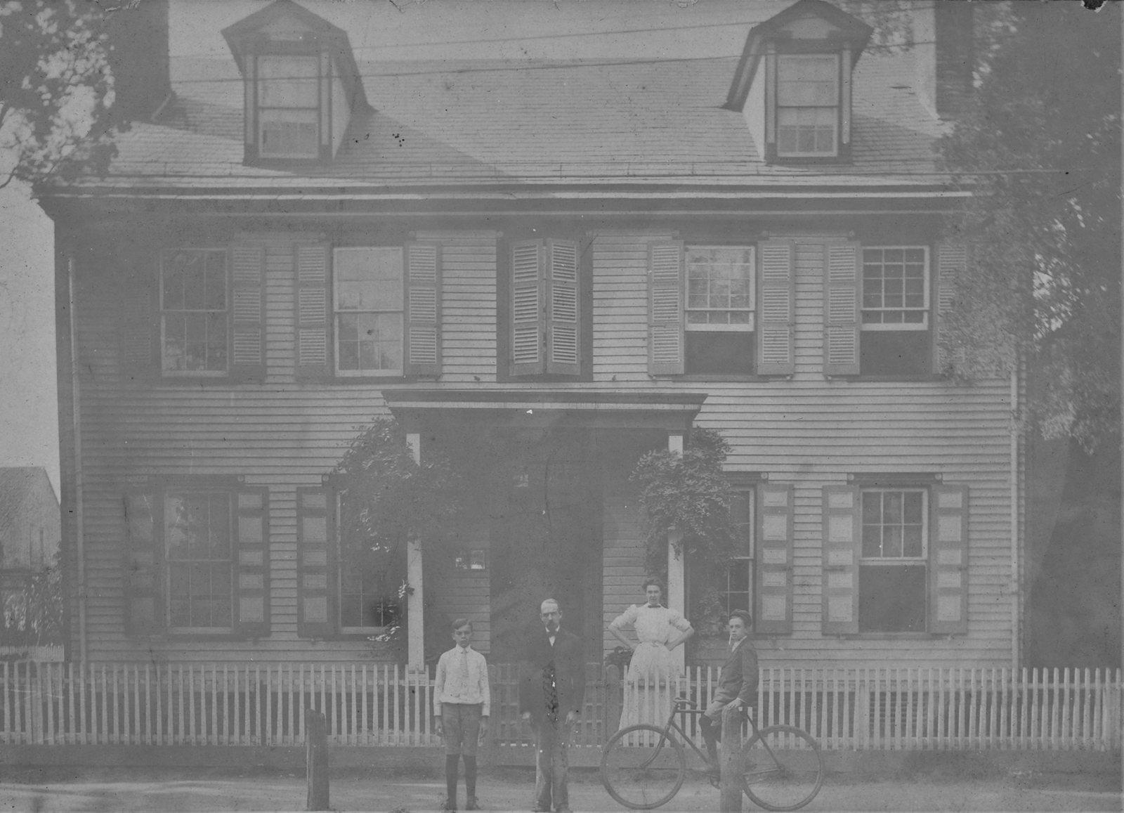 Four people and a bicycle at the picket fence of the tall frame house.