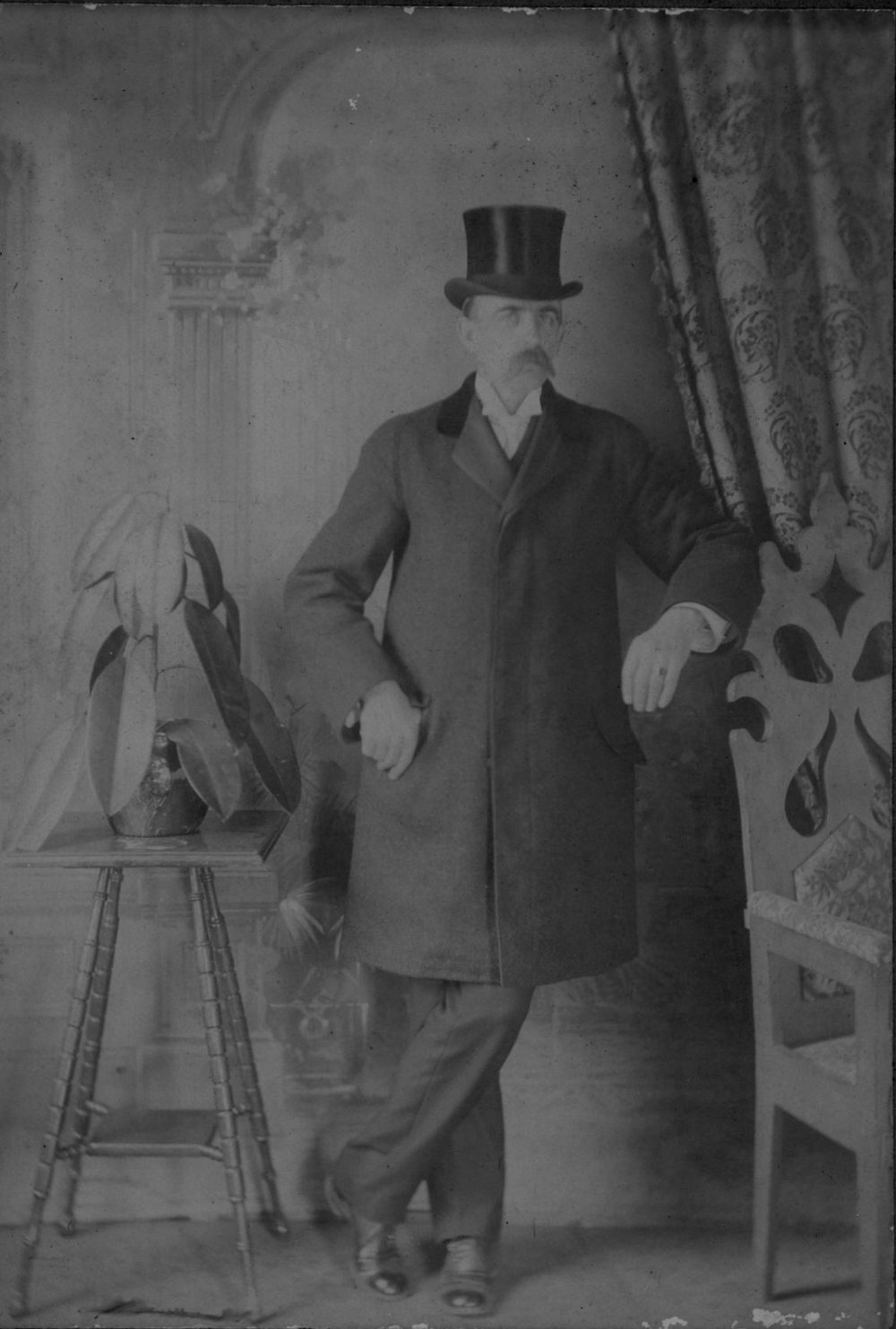 Studio portrait of a mustached man standing in a long coat and top hat, one arm resting on a chair.