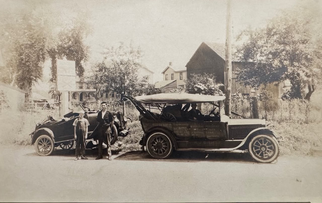 A man and a boy stand beside an early tow truck with hand-lettered service signs on its side, a wrecked open car hoisted on the hook behind it.